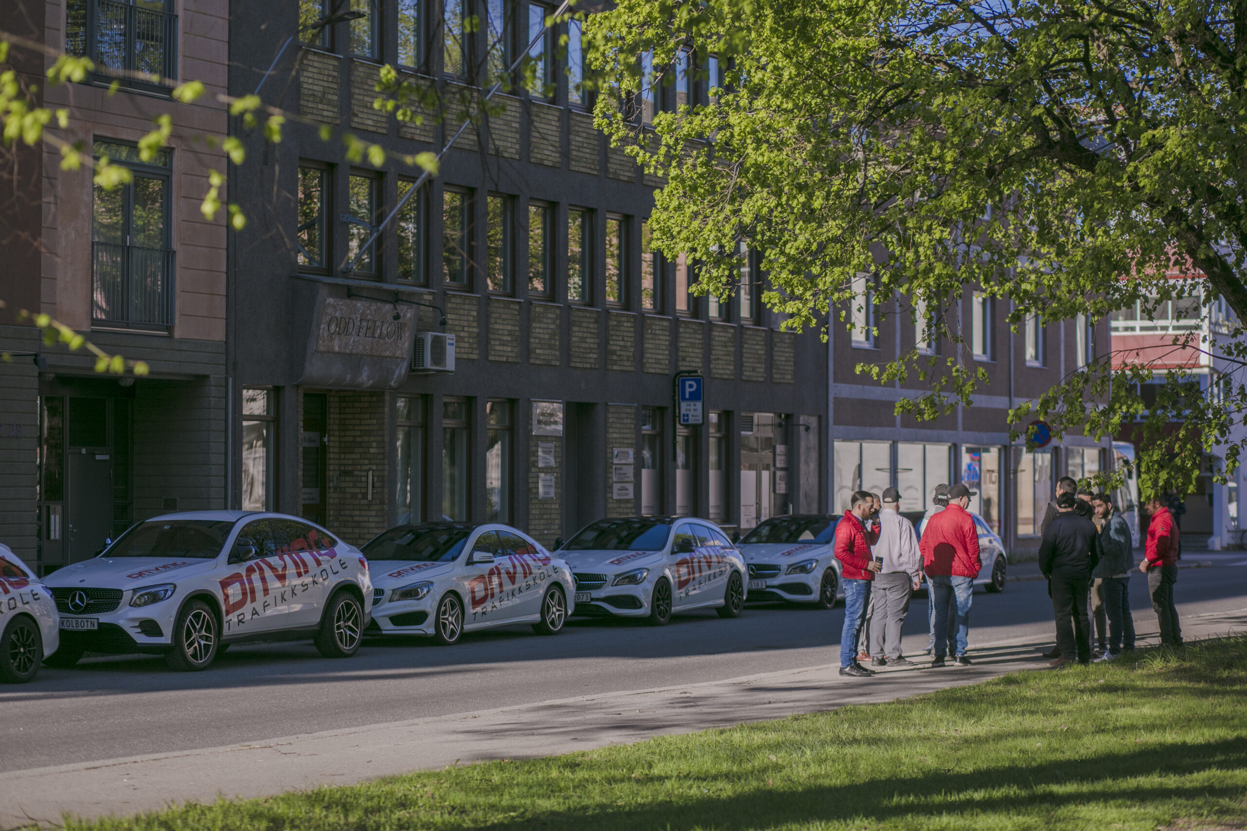Biler med Driving Trafikkskole sin logo står lukeparkert på rad utenfor en bygning. Kjørelærere med elever står på andre siden av veien og prater.