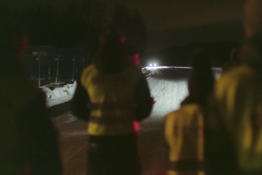 Et uskarpt bilde tatt i mørket utendørs, av kjøre- elever som står og ser på en bil med fjernlys i horisonten.